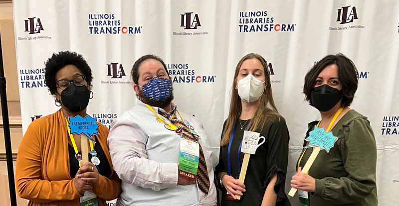 Michele Hunt, Sata Prescott, Alissa Droog, and Kimberly Shotick posing with library props in front of the Illinois Library Association Conference banner