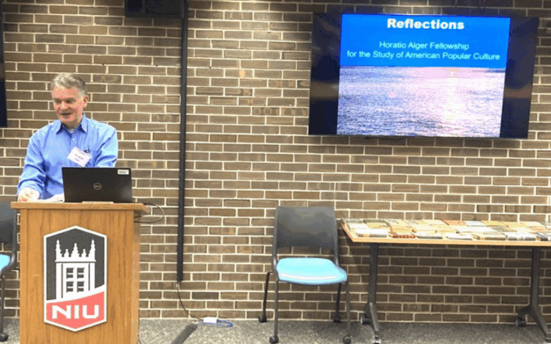 Fredrick Stark presenting at the Horatio Alger Conference in Founders Memorial Library, with a presentation slide titled 'Reflections: Horatio Alger Fellowship for the Study of American Popular Culture' on the screen behind him.