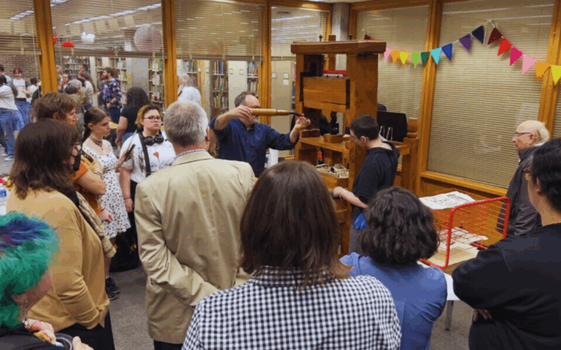 A crowd gathered around a demonstration at the grand opening of the NIU Book Lab. The image shows people observing a person operating an antique book press, with colorful paper garlands hanging in the background. Attendees of various ages and backgrounds are engaged, while some take photos or notes.