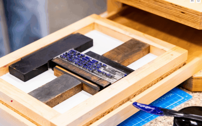 Close-up of a letterpress setup in the NIU BookLab, showing metal type locked into a wooden chase with wooden furniture and quoins. The type is inked with dark blue ink, and a spatula with ink rests nearby on a cutting mat.