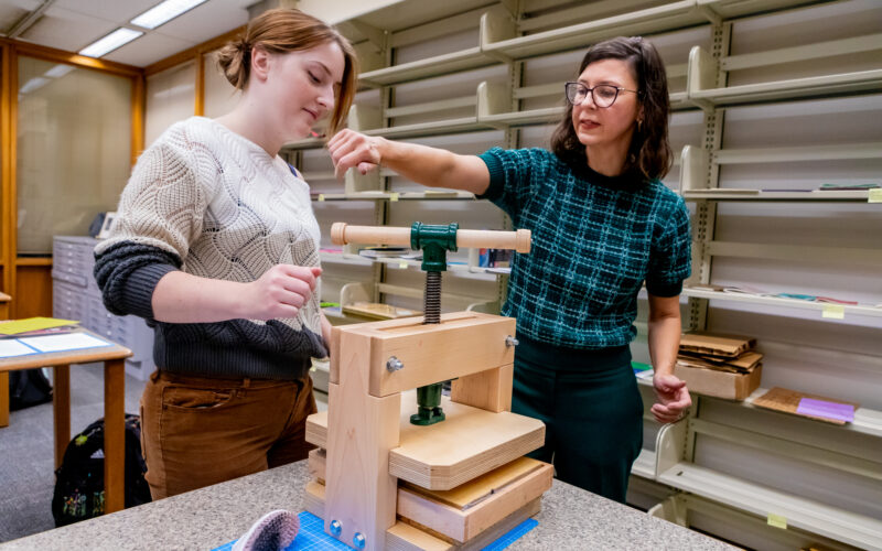 A librarian shows a student how to use a wooden book press on a worktable in a library conservation lab.