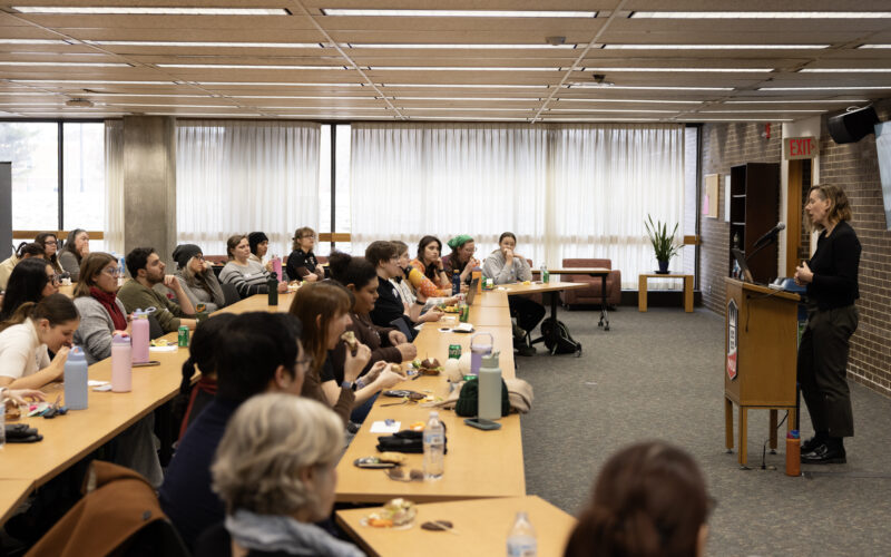 Audience seated at long tables listening to a speaker presenting at a podium in a classroom-style lecture room.