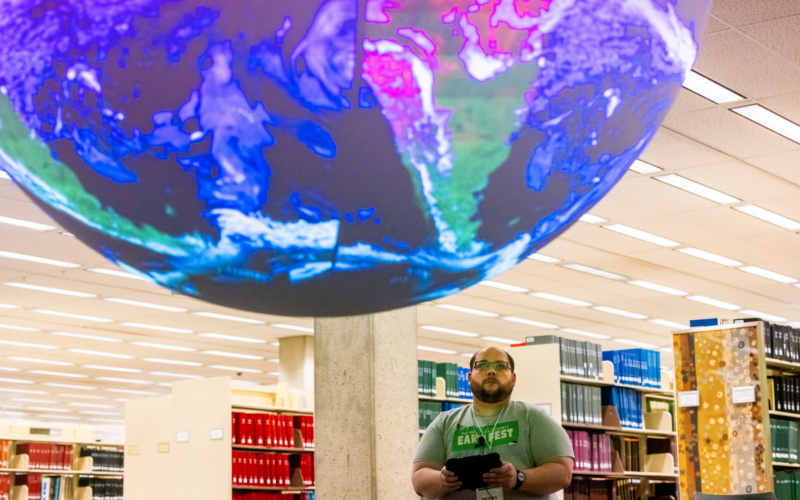 Person stands in a library looking up at a large suspended globe display above the stacks.