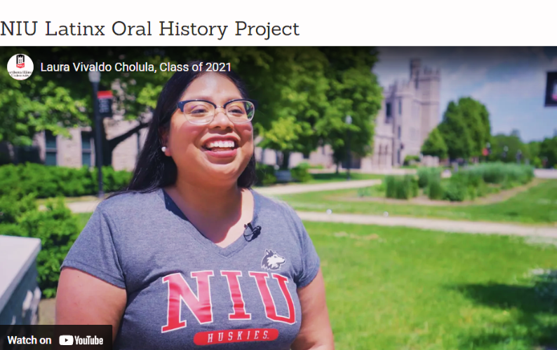 A woman wearing glasses and a gray NIU Huskies shirt smiles while standing on a sunny college campus lawn during an interview for the NIU Latinx Oral History Project.