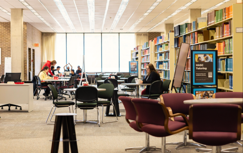 Students study and meet at tables inside a university library, with bookshelves and tutoring information visible nearby.
