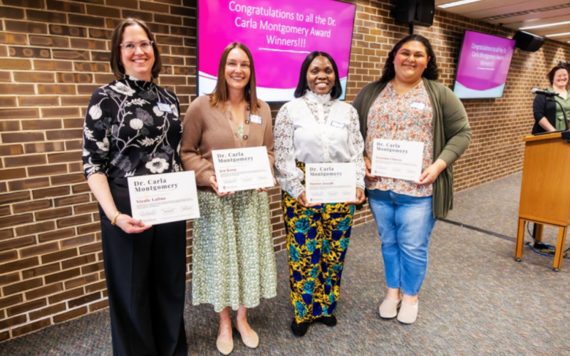 Four women stand side by side indoors, smiling and holding certificates for the Dr. Carla Montgomery Award, with a presentation screen behind them congratulating the winners.
