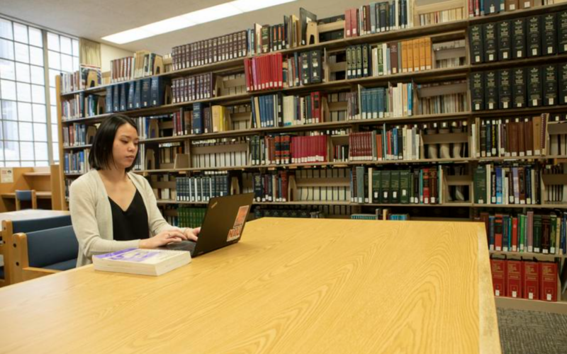Student working on a laptop at a large table in a library, with bookshelves filled with books in the background.