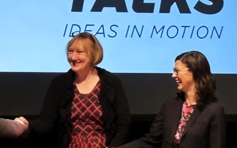 Two women smiling on stage at a conference, shaking hands in front of a screen displaying the words ‘Trek Talks: Ideas in Motion.’
